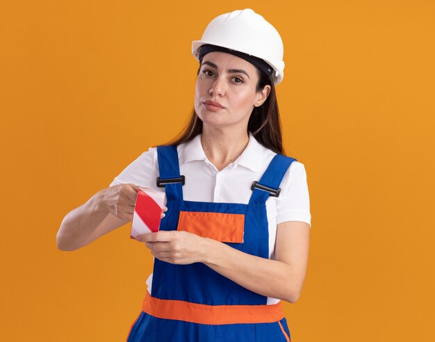 Confident young builder woman in uniform holding duct tape isolated on orange wall