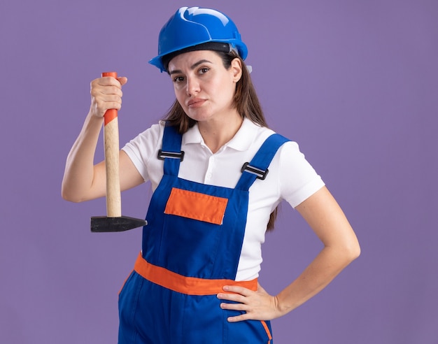 Confident young builder woman in uniform holding hammer putting hand on hip isolated on purple wall