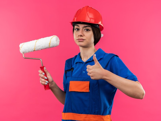 Confident young builder woman in uniform holding roller brush showing thumb up 