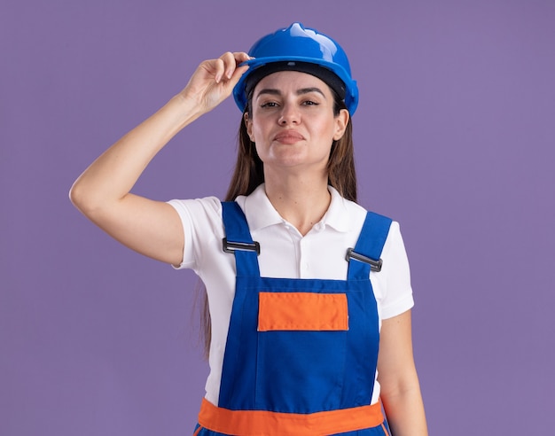 Free photo confident young builder woman in uniform holding safety helmet isolated on purple wall