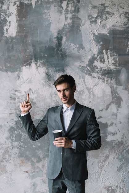 Free photo confident young businessman holding takeaway cup pointing finger upward against concrete wall