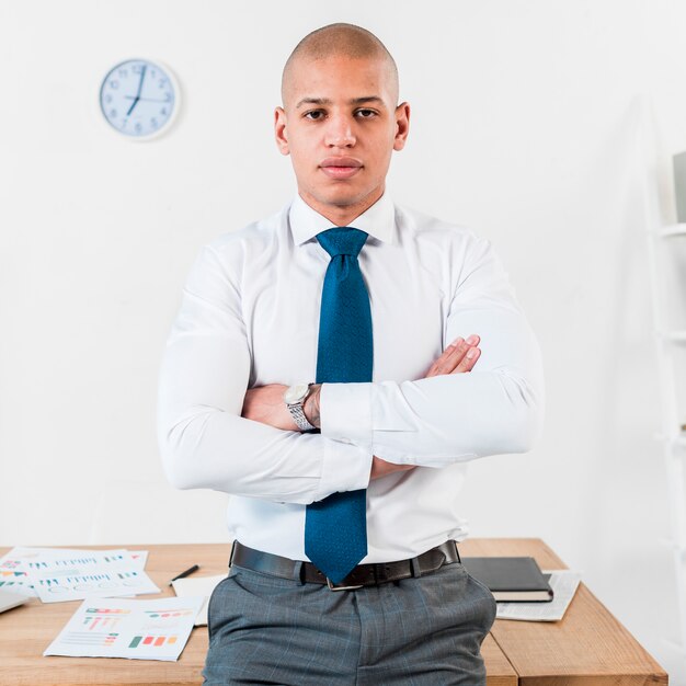 Confident young businessman standing in front of wooden table with his arm crossed