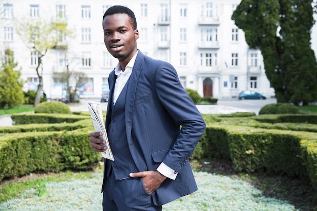 Confident young businessman with hands in his pocket holding newspaper looking at camera