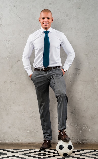 Free photo confident young businessman with his hands in the pocket and foot on soccer ball against grey concrete wall