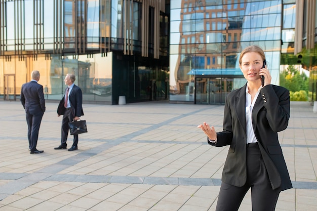 Confident young businesswoman in office suit talking on mobile phone and gesturing outdoors. Businesspeople and city building glass facade in background. Copy space. Business communication concept