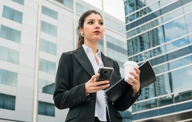 携帯電話を保持している本社ビルの外に立っている自信を持って若い実業家。使い捨てのコーヒーカップとフォルダーを手に