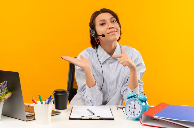 Confident young call center girl wearing headset sitting at desk showing empty hand and pointing at it isolated on orange 