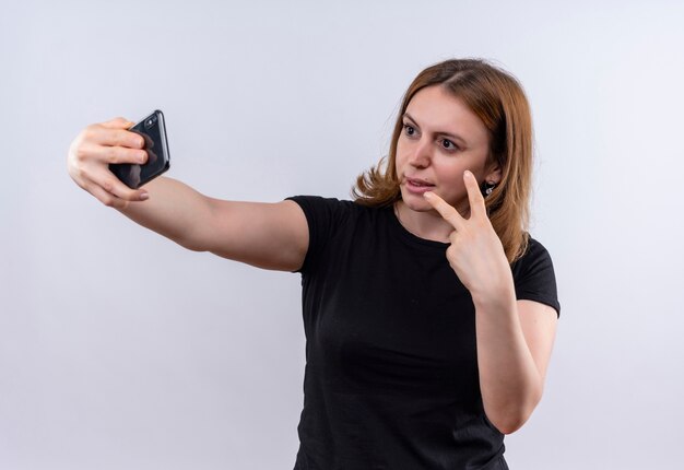 Confident young casual woman doing peace sign and taking selfie on isolated white space