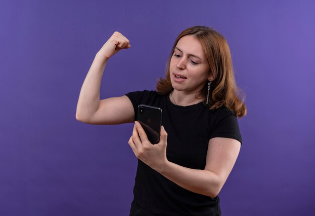 Confident young casual woman holding mobile phone and doing strong gesture looking at mobile phone on isolated purple space with copy space