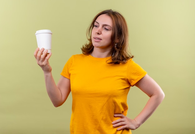 Confident young casual woman holding plastic coffee cup with hand on waist on isolated green space