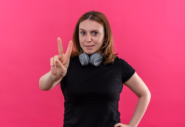 Confident young casual woman wearing headphones on neck doing peace sign on isolated pink space