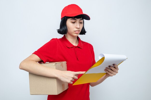 Confident young caucasian delivery girl holding cardboard box and looking at clipboard 