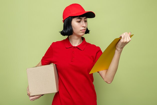 Confident young caucasian delivery girl holding cardboard box and looking at clipboard