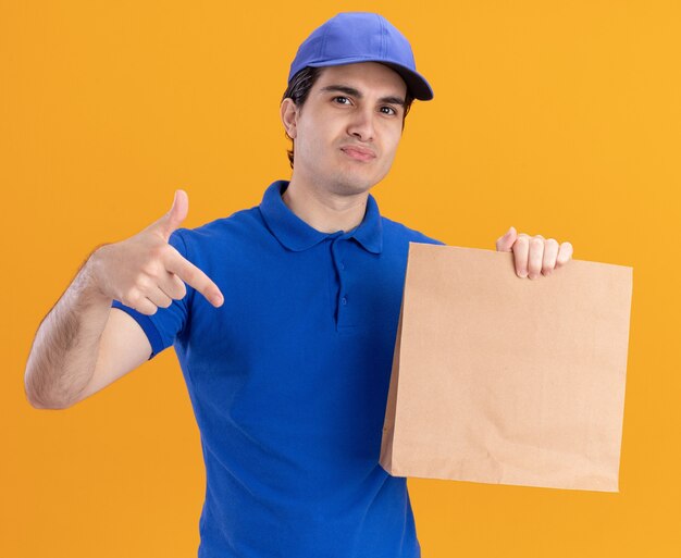 青い制服とオレンジ色の壁に分離された紙のパッケージを保持し、指しているキャップの自信を持って若い白人配達人