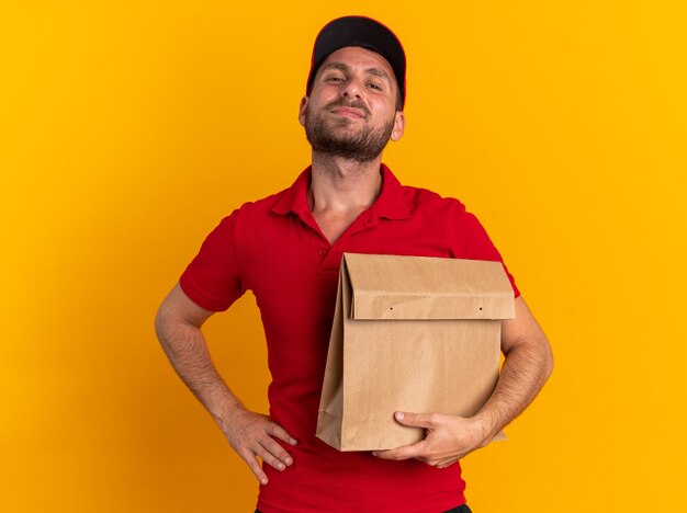 Confident young caucasian delivery man in red uniform and cap keeping hand on waist holding paper package looking at camera isolated on orange wall