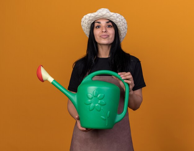 Confident young caucasian gardener girl wearing uniform and hat holding watering can 