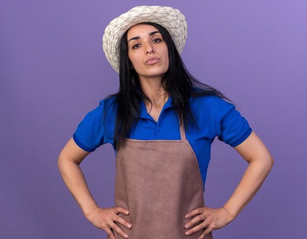 Уверенная молодая кавказская девушка-садовник в униформе и шляпе держит руки на талии, изолированной на фиолетовой стене