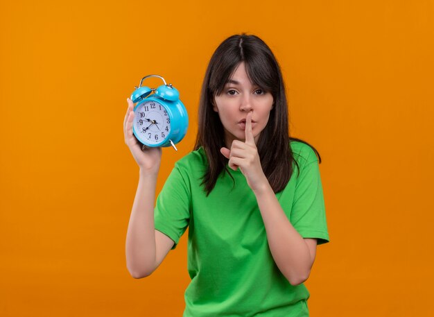 Confident young caucasian girl in green shirt holds clock and puts finger on mouth on isolated orange background