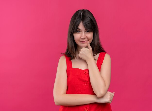 Confident young caucasian girl putting hand on chin and looking at camera on isolated pink background with copy space