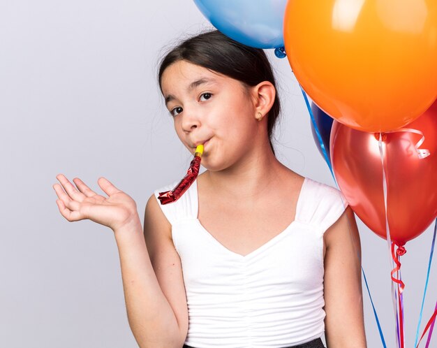 confident young caucasian girl standing with helium balloons blowing party whistle isolated on white wall with copy space