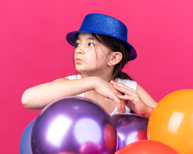 コピースペースでピンクの壁に隔離された側を見てヘリウム気球で立っている青いパーティーハットを身に着けている自信を持って若い白人の女の子