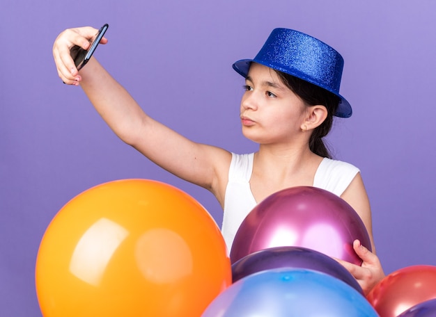 confident young caucasian girl wearing blue party hat taking selfie on phone standing with helium balloons isolated on purple wall with copy space