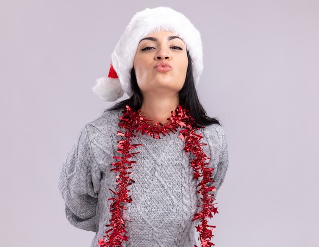 Уверенная молодая кавказская девушка в новогодней шапке и гирлянде из мишуры на шее, держа руки за спиной, глядя в камеру, делая жест поцелуя на белом фоне с копией пространства