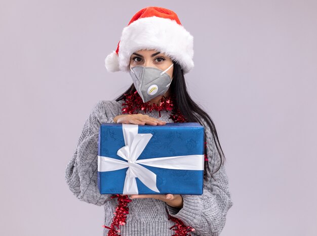 Confident young caucasian girl wearing christmas hat and tinsel garland around neck with protective mask looking at camera holding gift package isolated on white background with copy space