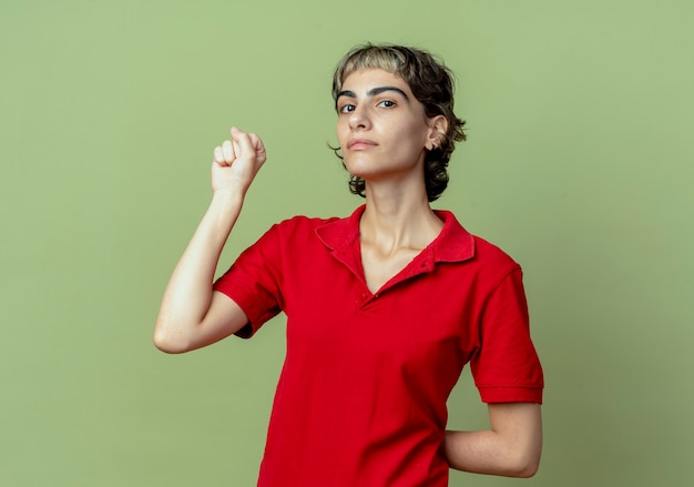 Confident young caucasian girl with pixie haircut clenching fist and hiding another hand behind back isolated on olive green background with copy space