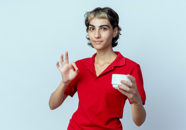 Уверенная молодая кавказская девушка с прической пикси держит чашку и делает знак ОК, изолированные на белом фоне с копией пространства
