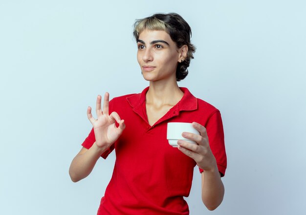 Confident young caucasian girl with pixie haircut holding cup doing ok sign and looking at side isolated on white background with copy space