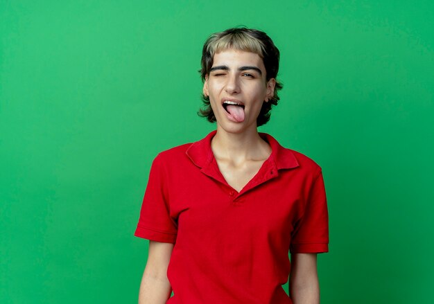 Confident young caucasian girl with pixie haircut winking and showing tongue isolated on green background with copy space