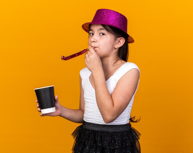 confident young caucasian girl with purple party hat holding paper cup and blowing party whistle isolated on orange wall with copy space