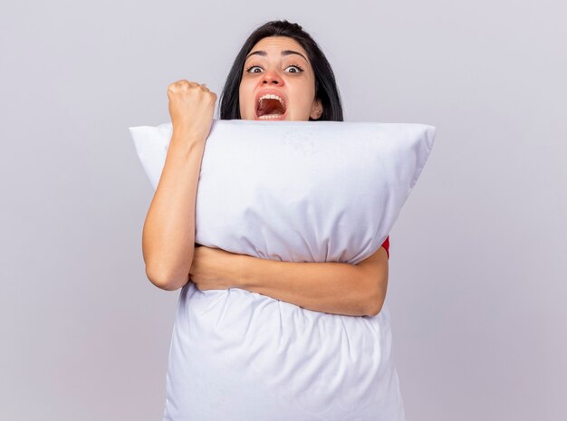Confident young caucasian ill girl hugging pillow looking at camera from behind it doing be strong gesture isolated on white background with copy space