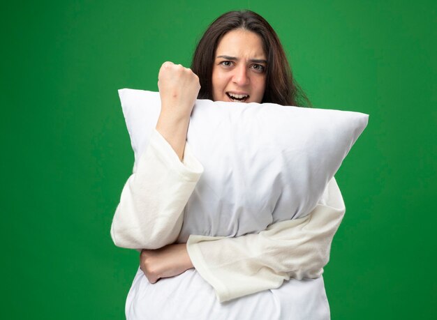 Confident young caucasian ill girl wearing robe hugging pillow looking at camera doing be strong gesture isolated on green background with copy space