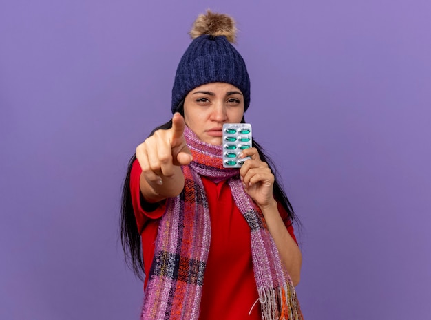 Free photo confident young caucasian ill girl wearing winter hat and scarf holding pack of capsules looking and pointing  isolated on purple wall with copy space