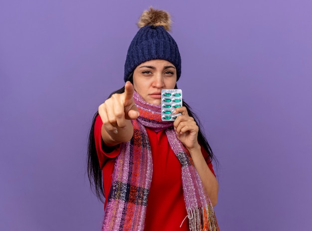 Confident young caucasian ill girl wearing winter hat and scarf holding pack of capsules looking and pointing  isolated on purple wall with copy space