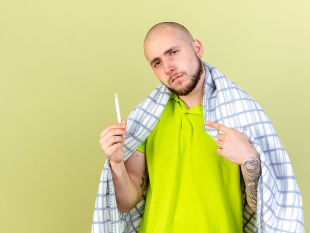 格子縞のホールドに包まれた自信を持って若い白人の病気の男は、コピースペースのあるオリーブグリーンの壁に隔離された温度計を指しています