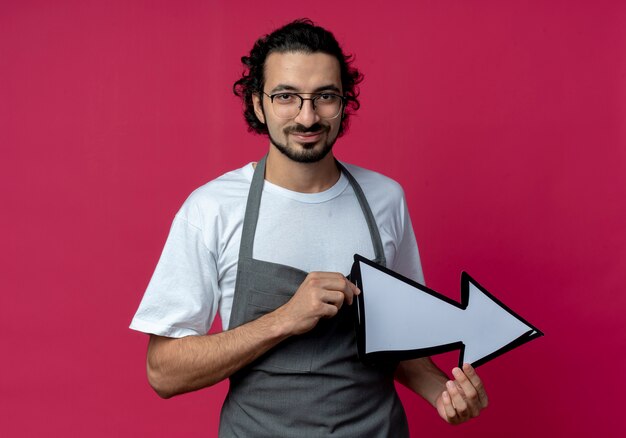 Confident young caucasian male barber wearing glasses and wavy hair band in uniform holding arrow mark which is pointing at side isolated on crimson background