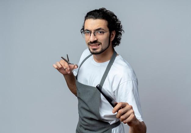Confident young caucasian male barber wearing glasses and wavy hair band in uniform standing in profile view holding scissors and stretching out comb