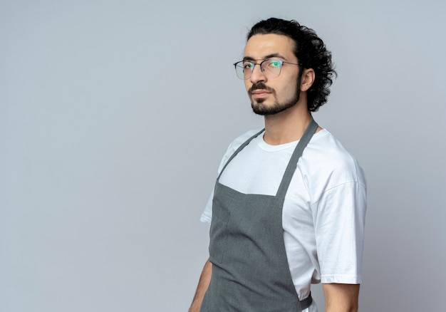 Уверенный молодой кавказский мужчина-парикмахер в очках и волнистой повязке для волос в униформе, стоя в профиль, глядя в камеру, изолированную на белом фоне с копией пространства
