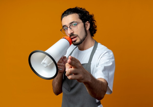 Confident young caucasian male barber wearing uniform and glasses talking by speaker and pointing at camera and looking at camera isolated on orange background with copy space