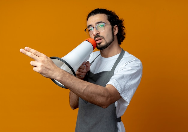 Уверенный молодой кавказский мужчина-парикмахер в униформе и очках разговаривает спикером и показывает пальцами прямо и смотрит в камеру, изолированную на оранжевом фоне с копией пространства