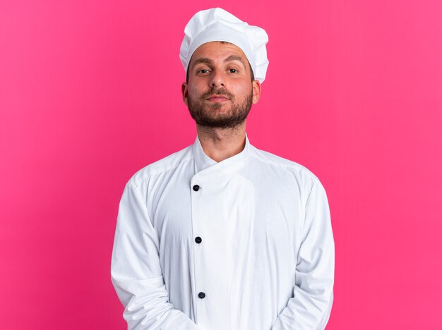 Confident young caucasian male cook in chef uniform and cap keeping hands together looking at camera isolated on pink wall