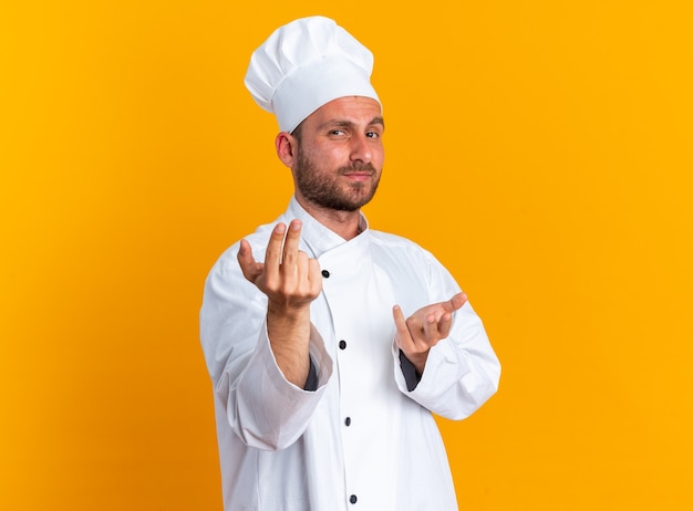 자신감 있는 젊은 백인 남성 요리사 유니폼을 입고 카메라를 쳐다보는 모자가 주황색 벽에 격리된 몸짓으로 여기에 온다