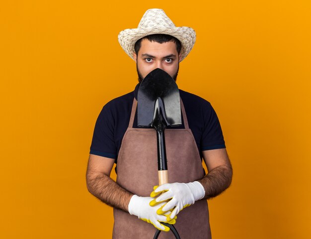 confident young caucasian male gardener wearing gardening hat and gloves holding spade isolated on orange wall with copy space