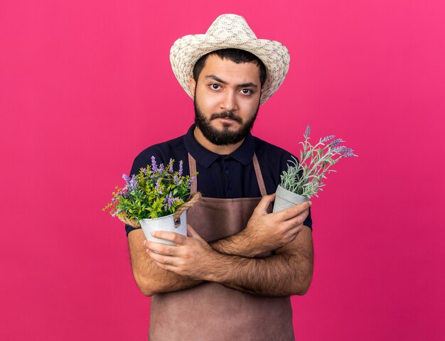 confident young caucasian male gardener wearing gardening hat holding flowerpots crossing arms isolated on pink wall with copy space