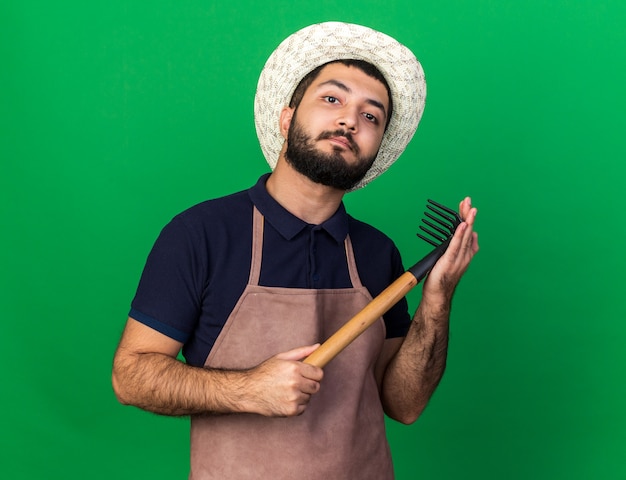 confident young caucasian male gardener wearing gardening hat holding rake and looking  isolated on green wall with copy space