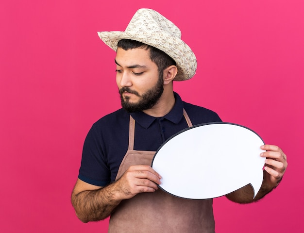 confident young caucasian male gardener wearing gardening hat holding speech balloon and looking at side isolated on pink wall with copy space