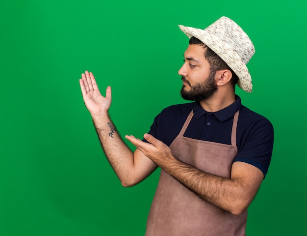 confident young caucasian male gardener wearing gardening hat looking and pointing at his empty hand isolated on green wall with copy space